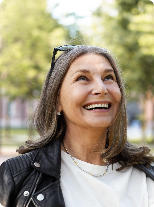 Newcastle Specialist Prosthodontics Smiling woman with long gray hair looking up while standing outdoors in a park.